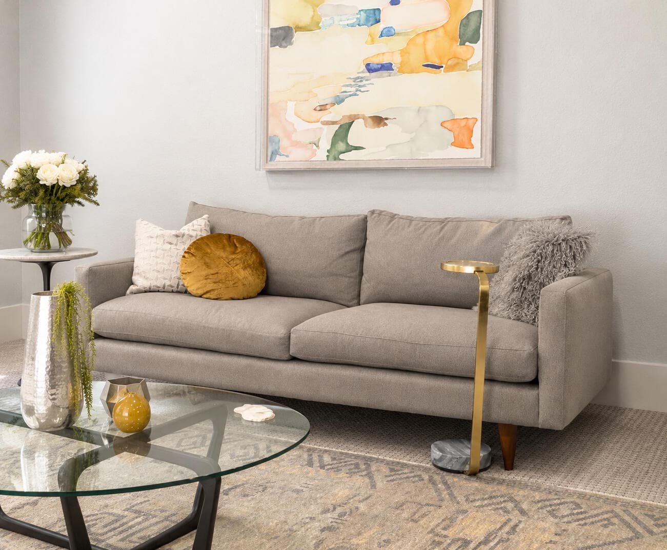 Grey sofa with a mustard cushion, a glass coffee table and a brass side table under a watercolour