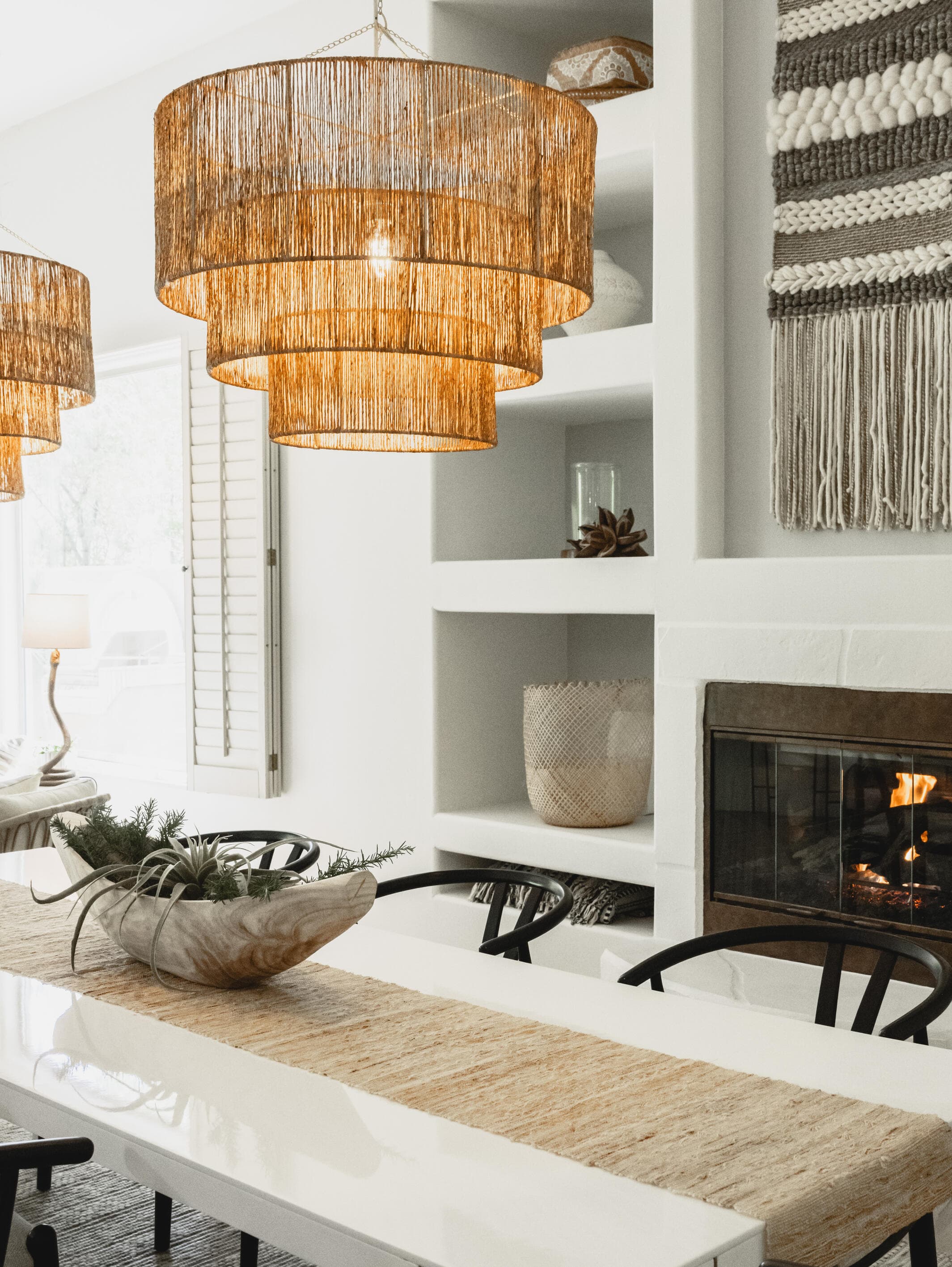 Dining table under tiered jute pendants, with black wishbone chairs and white plaster shelves